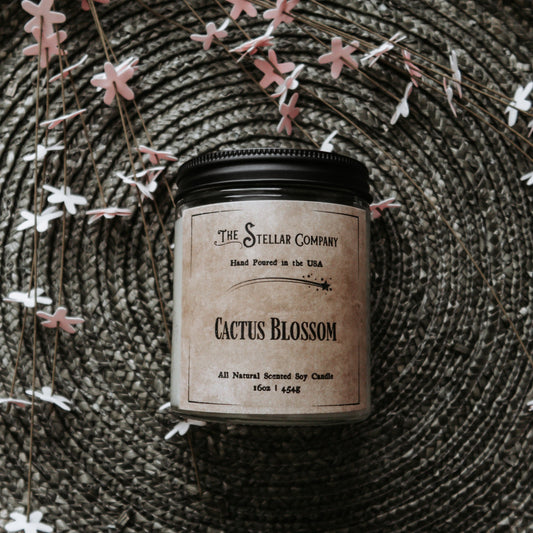 The Stellar Company Cactus Blossom scented soy candle, hand-poured in the USA, displayed on a woven mat with delicate flower-shaped accents. This all-natural, 16oz candle offers a refreshing floral fragrance, perfect for home decor or gifting